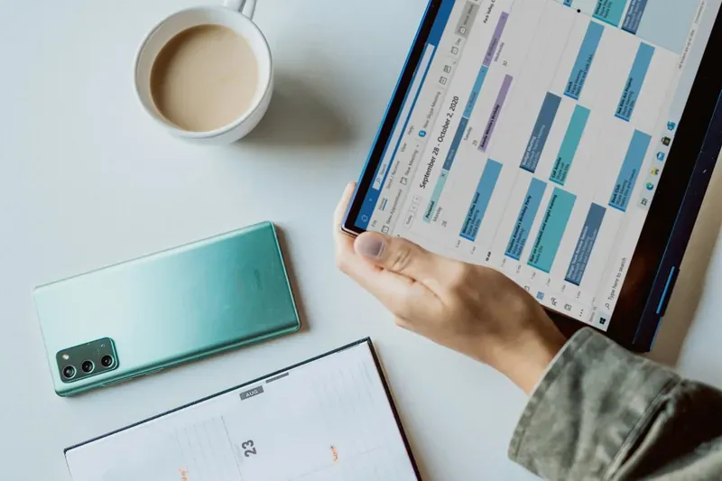 Mano che tiene un tablet con il calendario della settimana, accanto a un caffè e a un telefono