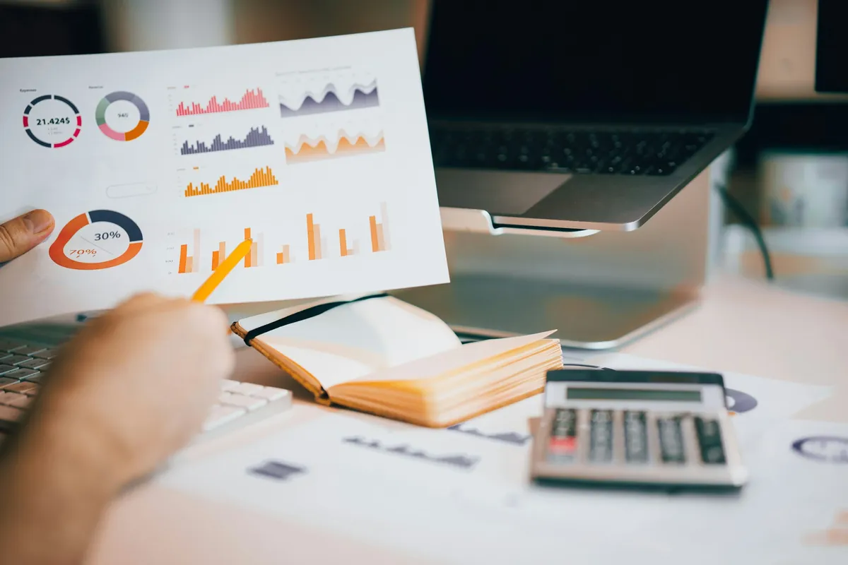 Mano che indica con una matita i grafici di un report stampato, accanto a un laptop e a una calcolatrice